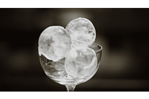 A high-contrast, black and white image of three perfectly clear, spherical Hoshizaki ice balls stacked in an elegant, stemmed cocktail glass for a premium beverage presentation