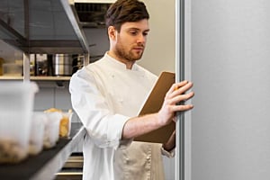 A chef in a white uniform reviews a clipboard, taking inventory next to a stainless steel commercial freezer or refrigerator from Hoshizaki in a kitchen setting