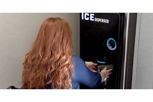 A hotel guest with long red hair uses the self-service "Push for Ice" button on a black, wall-mounted Hoshizaki ice dispenser