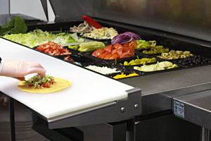 A close-up of a worker's hand assembling a taco or wrap on a cutting board attached to a Hoshizaki Mega-Tops refrigerated prep table filled with fresh ingredients