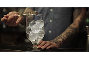 A bartender with tattooed arms stirring a cocktail in a glass filled with clear Hoshizaki Sphere Ice