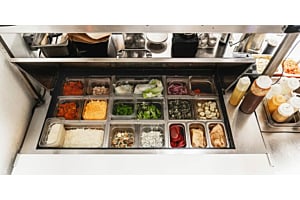 Hoshizaki refrigerated prep table display well stocked with fresh ingredients for food assembly in a fast-casual setting