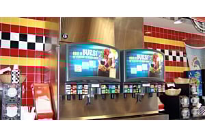 Two Hoshizaki twinpour chewable ice and beverage dispensers are featured against a vibrant yellow, red, and black tiled wall in a quick-service restaurant or convenience store