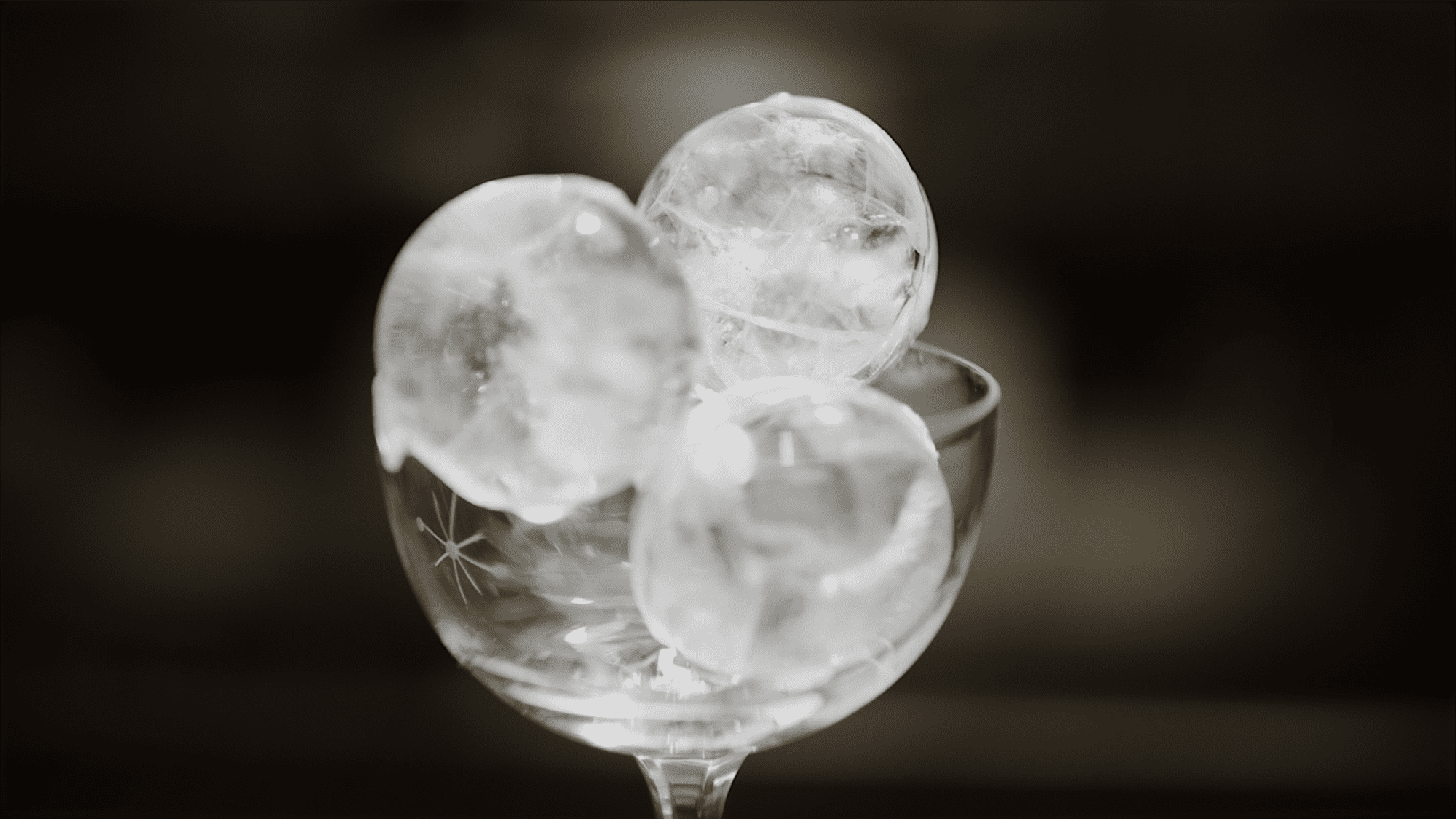A high-contrast, black and white image of three perfectly clear, spherical Hoshizaki ice balls stacked in an elegant, stemmed cocktail glass for a premium beverage presentation
