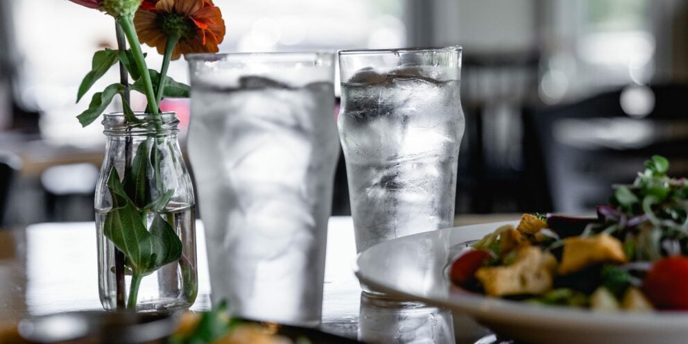 Two tall glasses of water with Hoshizaki Crescent Ice on a restaurant table next to a floral centerpiece and food