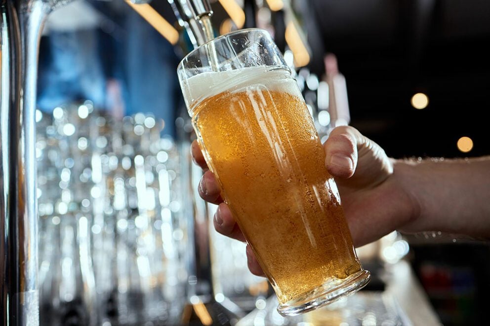 A hand holds a glass of golden beer being freshly poured from a chrome tap, demonstrating the use of Hoshizaki direct draw systems