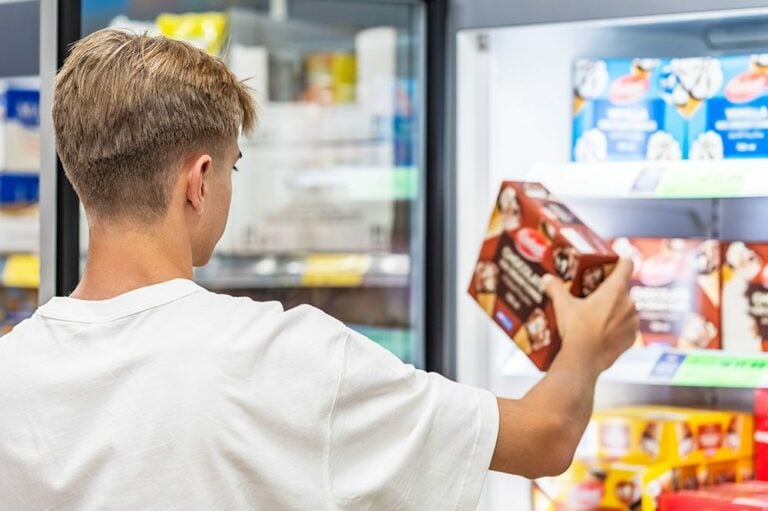 A customer holds a box of frozen treats while looking into a brightly lit Hoshizaki glass door merchandiser freezer in a retail environment