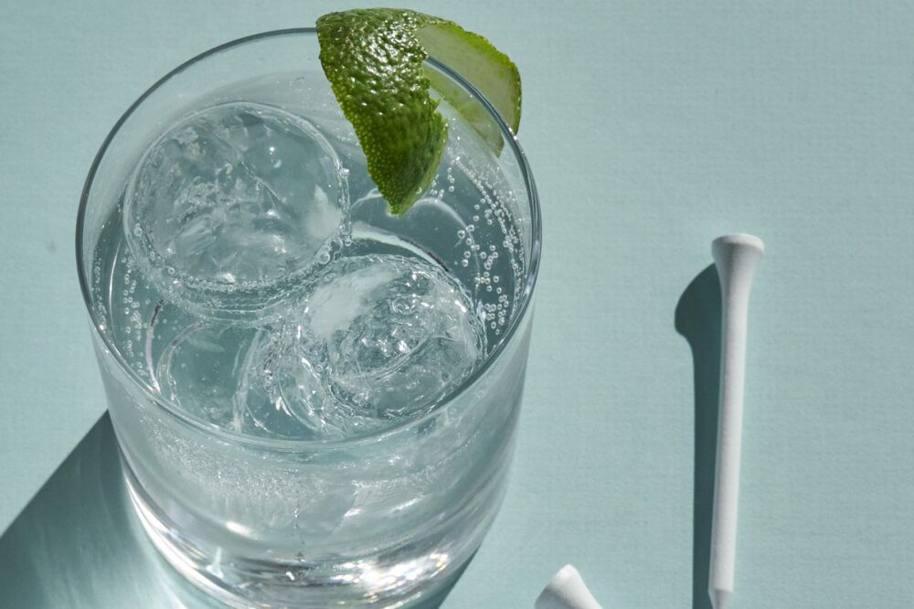 A clear beverage garnished with a lime and featuring two perfectly clear Hoshizaki sphere ice balls, with a white golf tee visible on the light blue surface