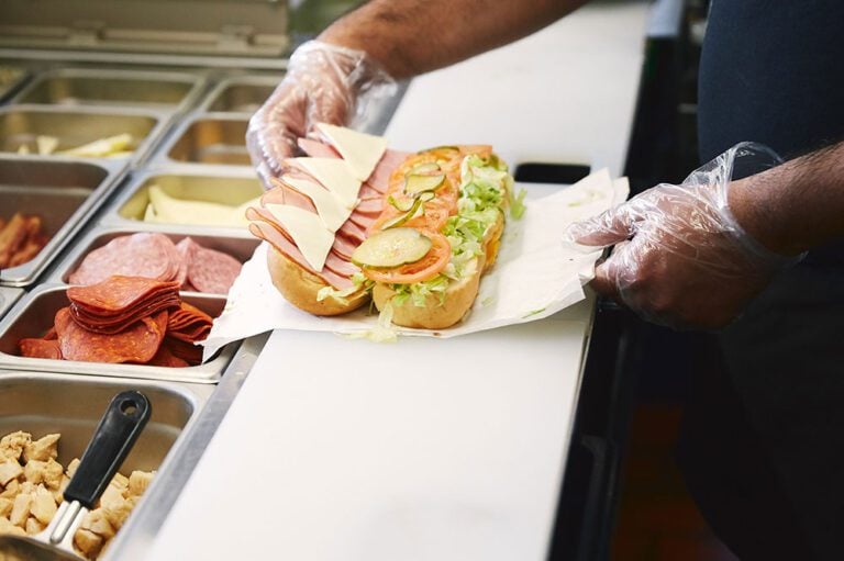 A food service worker, wearing gloves, layers meats and cheeses onto a sandwich, utilizing a Hoshizaki Valiance refrigerated prep table to keep ingredients fresh